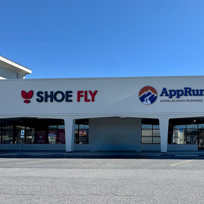 A storefront with SHOE FLY in red and black letters and a butterfly logo, next to AppRun Appalachian Running Company with a mountain logo, both under a clear blue sky.