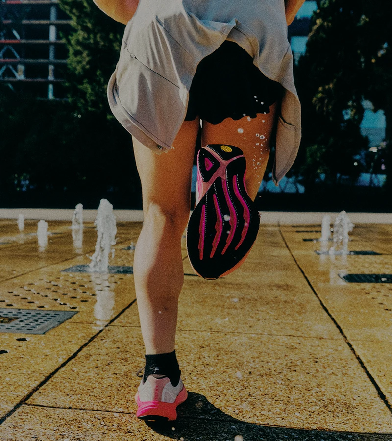 A person in athletic wear runs through a plaza with water jets shooting up from the ground, droplets splashing around their legs and shoes.