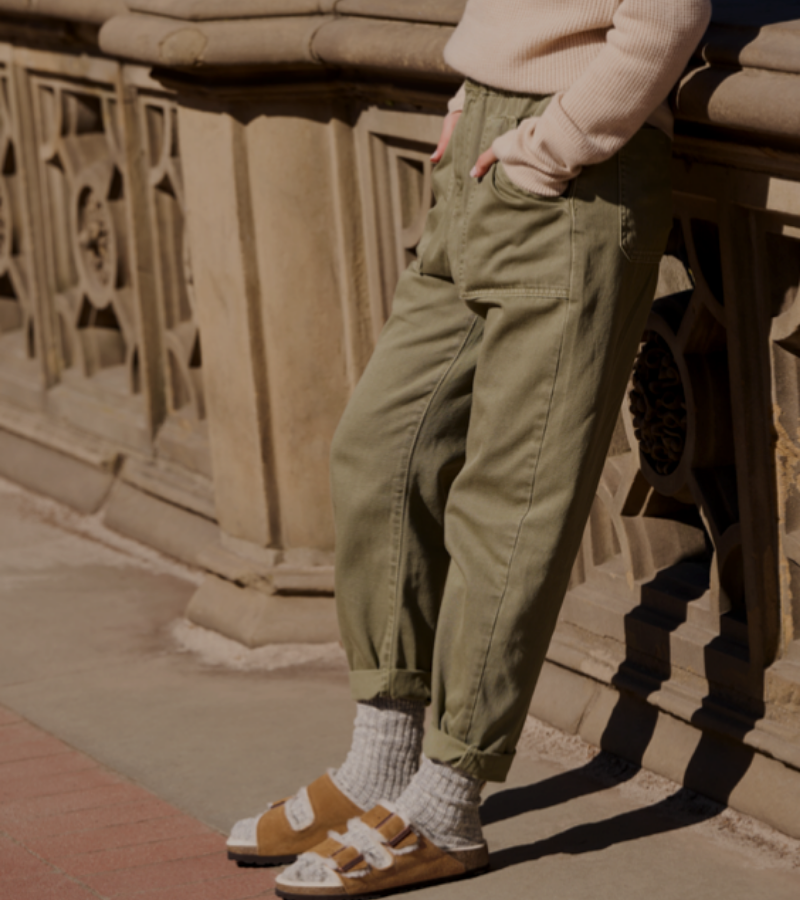 A person stands outdoors by a stone railing, wearing rolled-up green pants, a light sweater, socks, and brown sandals. Their hands are in their pockets and their head is out of frame.