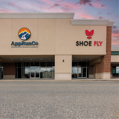 Storefronts of AppRunCo (Appalachian Running Company) and SHOEFly, side by side in a strip mall at dusk, with empty parking lot in front.