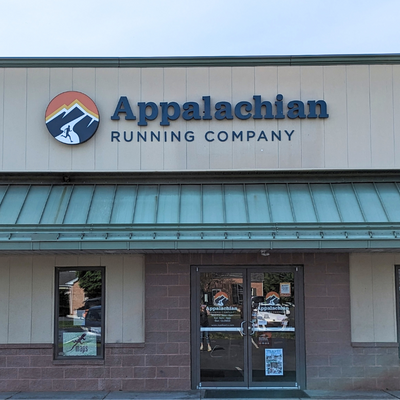 Storefront for Appalachian Running Company, with a mountain logo above the entrance, a green metal awning, and glass doors reflecting the street and trees. The sign reads Appalachian Running Company..