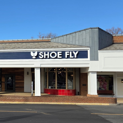 A storefront with a navy sign that reads SHOE FLY in white letters, featuring a small white fly graphic above the text. The building has large windows and a red counter visible inside.