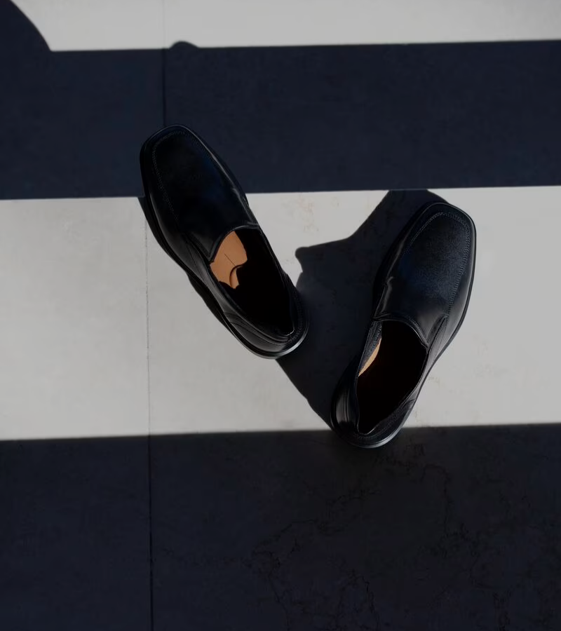 A pair of black slip-on shoes placed on a tiled floor with alternating light and shadow stripes across the surface.