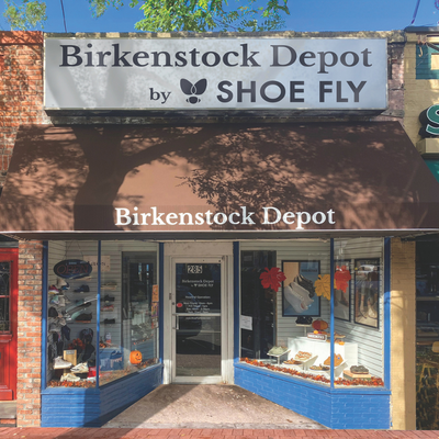 Storefront of Birkenstock Depot by Shoe Fly with brown awning, white sign, and shoe displays in windows decorated with fall leaves and a pumpkin. Reflective glass shows trees and sky.