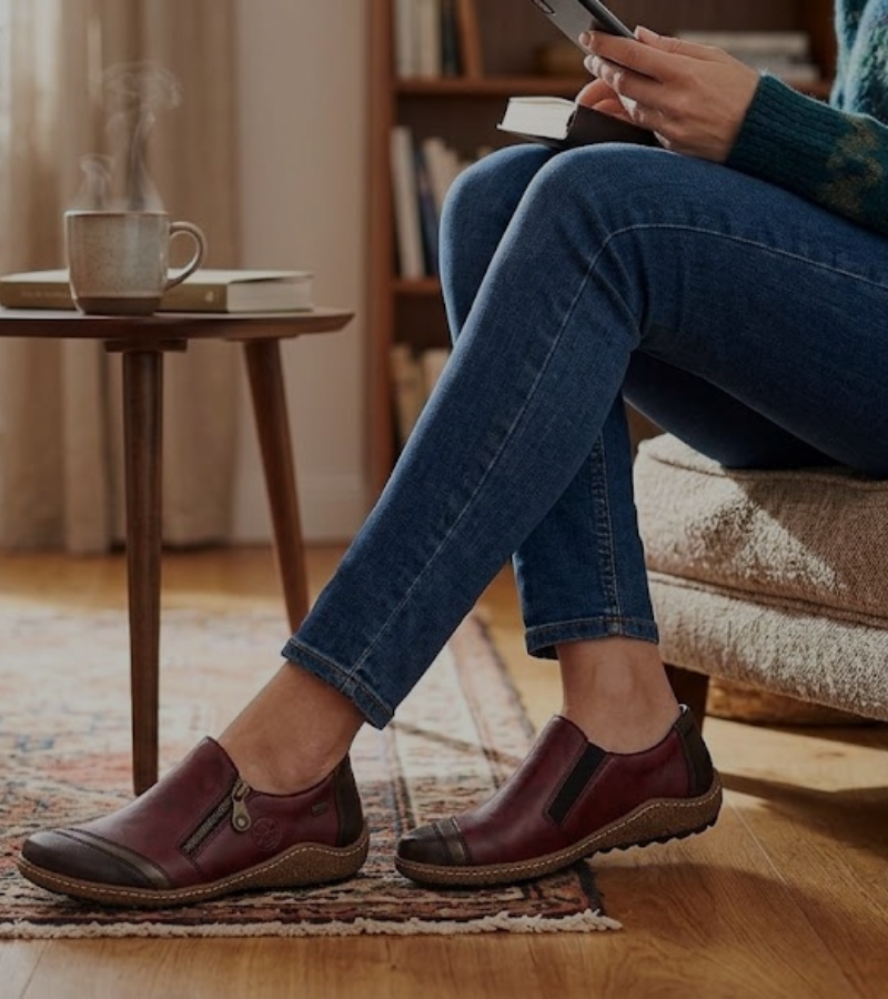 A person in jeans and red slip-on shoes sits on a couch, holding a phone and notebook. A steaming mug sits on a small table nearby, with a bookshelf in the background.