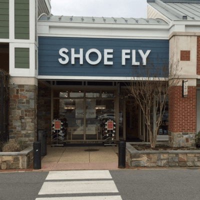 Storefront of a shop called Shoe Fly with large glass windows, a stone and brick exterior, and two sale signs by the entrance.