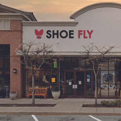 Storefront with a sign reading SHOE FLY in bold letters. The entrance is flanked by leafless trees, potted plants, and a wooden bench. The sky shows early evening light.
