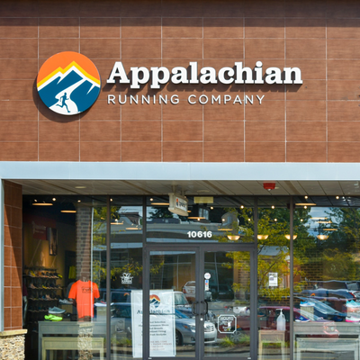 Storefront of Appalachian Running Company with large sign and mountain logo above glass doors. Reflections of cars and trees are visible in the windows. Athletic gear and shoes are displayed inside.