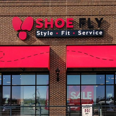 Storefront with a red awning and a sign that reads SHOE FLY and Style • Fit • Service. A large red fly logo is above the text. A sale sign in the window advertises 50% off apparel.