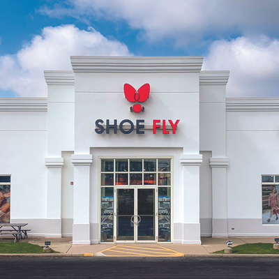 A white storefront with glass doors displays the sign SHOE FLY in black and red letters, with a red butterfly logo above. Theres a picnic table to the left and posters in the windows.
